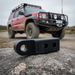 The Sherpa 4x4 Tow Hitch - Grey by Sherpa 4x4 sits on a rock in focus, showcasing this must-have offroad accessory; behind it, a red off-road vehicle with roof rack and bull bar is blurred on a dirt road.
