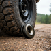 A rugged off-road vehicle tire sits on gravel with the Sherpa 4x4 Rope Recovery Ring 10T winch pulley positioned beneath its edge. The black Sherpa 4x4 accessory contrasts against the ground, with blurred forest scenery in the background.