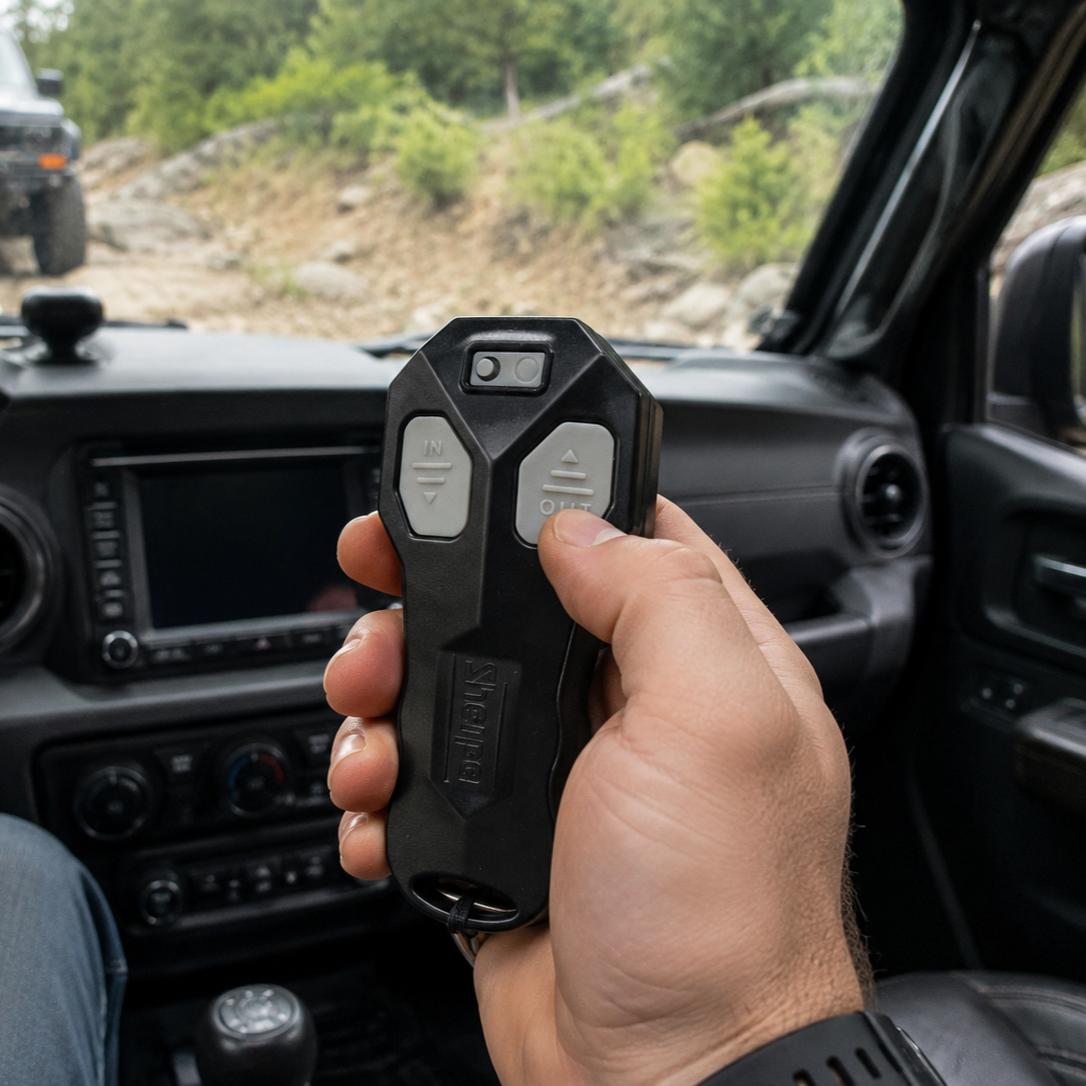 A hand uses the Sherpa 4x4 Wireless Receiver & Remote by Sherpa 4x4 to operate a winch on an off-road vehicle stuck in mud on a forest trail, surrounded by trees and dirt.