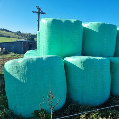 Large green hay bales wrapped in Paddock Silage Bale Wrap Film are stacked outdoors on a sunny day, with a grassy field, wooden shed, and power pole in the background.