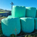 Large green hay bales wrapped in Paddock Silage Bale Wrap Film are stacked outdoors on a sunny day, with a grassy field, wooden shed, and power pole in the background.
