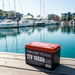 A Trolltek 12V Trolltek™ LiFePO4 Lithium Battery with Bluetooth sits on a wooden dock by a marina, boats and modern buildings reflecting in the calm water behind it.
