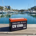 A 12V Trolltek™ LiFePO4 Lithium Battery with Bluetooth sits on a dock, with sailboats and modern waterfront buildings in the background on a sunny day.
