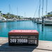 A Trolltek 24V Trolltek™ LiFePO4 Lithium Battery with Bluetooth rests on a wooden dock in a marina, surrounded by yachts and palm trees reflected in the calm water beneath a clear sky.