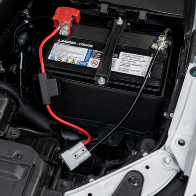 Close-up of a Trolltek™ Fused Battery Connector Cable - 50A by Trolltek installed under the hood, with red and black cables securely connected to the terminals. The heavy-duty cable setup is ideal for dual battery systems, and visible on the battery are warning labels and safety instructions.