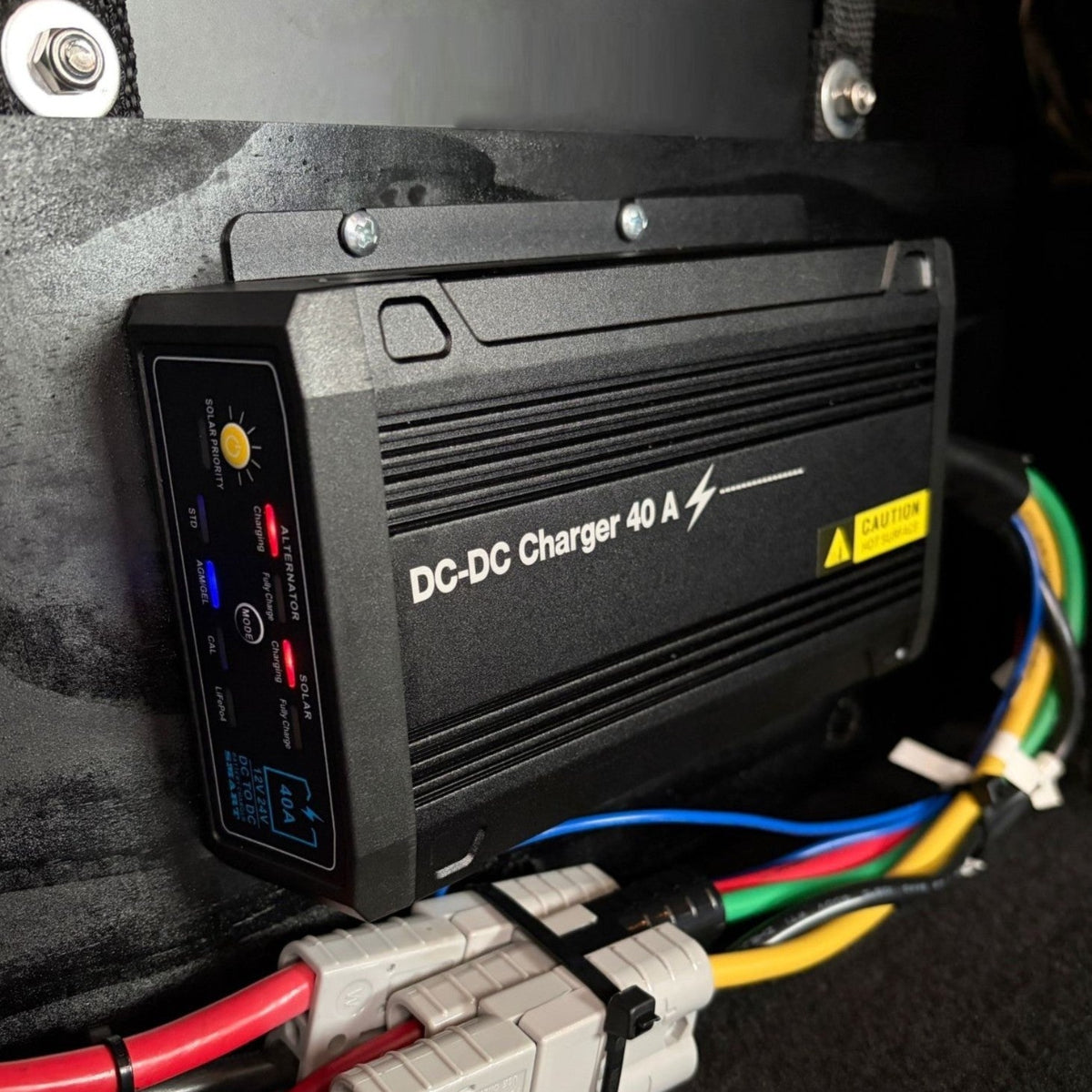 A Trolltek™ 40Amp DC/DC Charger with MPPT Solar Input by Trolltek in black is mounted on a wall. Colorful cables—blue, red, yellow, green, and black—connect at the bottom of the device. A prominent yellow caution sticker is visible. Designed as a reliable DC to DC Lithium Battery Charger for versatile applications, it also serves efficiently as an MPPT Solar Regulator for optimal performance.