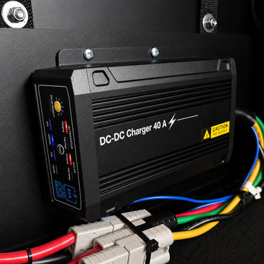 A Trolltek™ 40Amp DC/DC Charger with MPPT Solar Input by Trolltek in black is mounted on a wall. Colorful cables—blue, red, yellow, green, and black—connect at the bottom of the device. A prominent yellow caution sticker is visible. Designed as a reliable DC to DC Lithium Battery Charger for versatile applications, it also serves efficiently as an MPPT Solar Regulator for optimal performance.