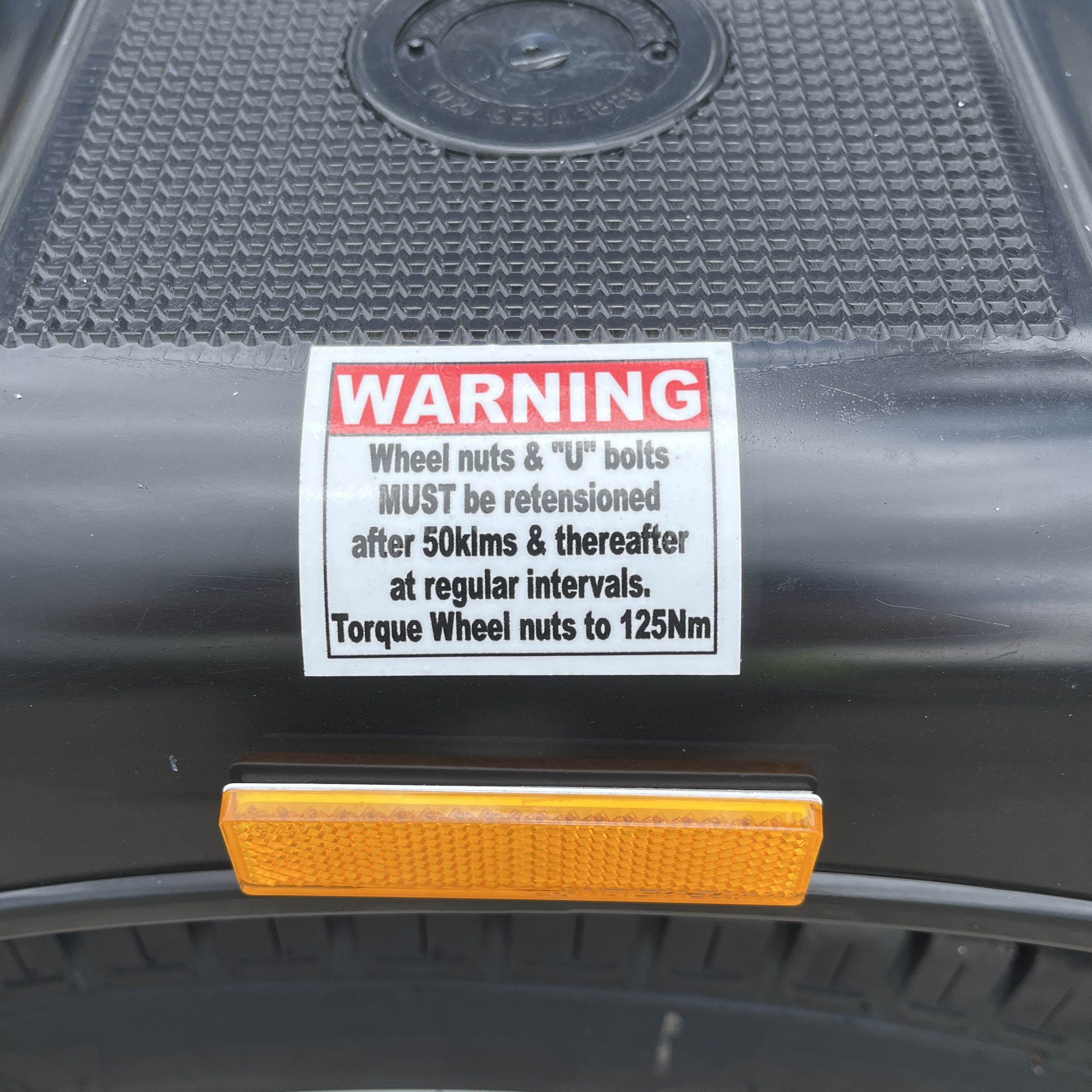 A close-up of a warning label on a black part of the Paddock Mini Loader & Excavator Alloy Equipment Trailer by Paddock, instructing to retension wheel nuts and U bolts after 50km and regularly, torque nuts to 125Nm; a yellow reflector sits below.