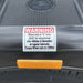 A close-up of a warning label on a black part of the Paddock Mini Loader & Excavator Alloy Equipment Trailer by Paddock, instructing to retension wheel nuts and U bolts after 50km and regularly, torque nuts to 125Nm; a yellow reflector sits below.