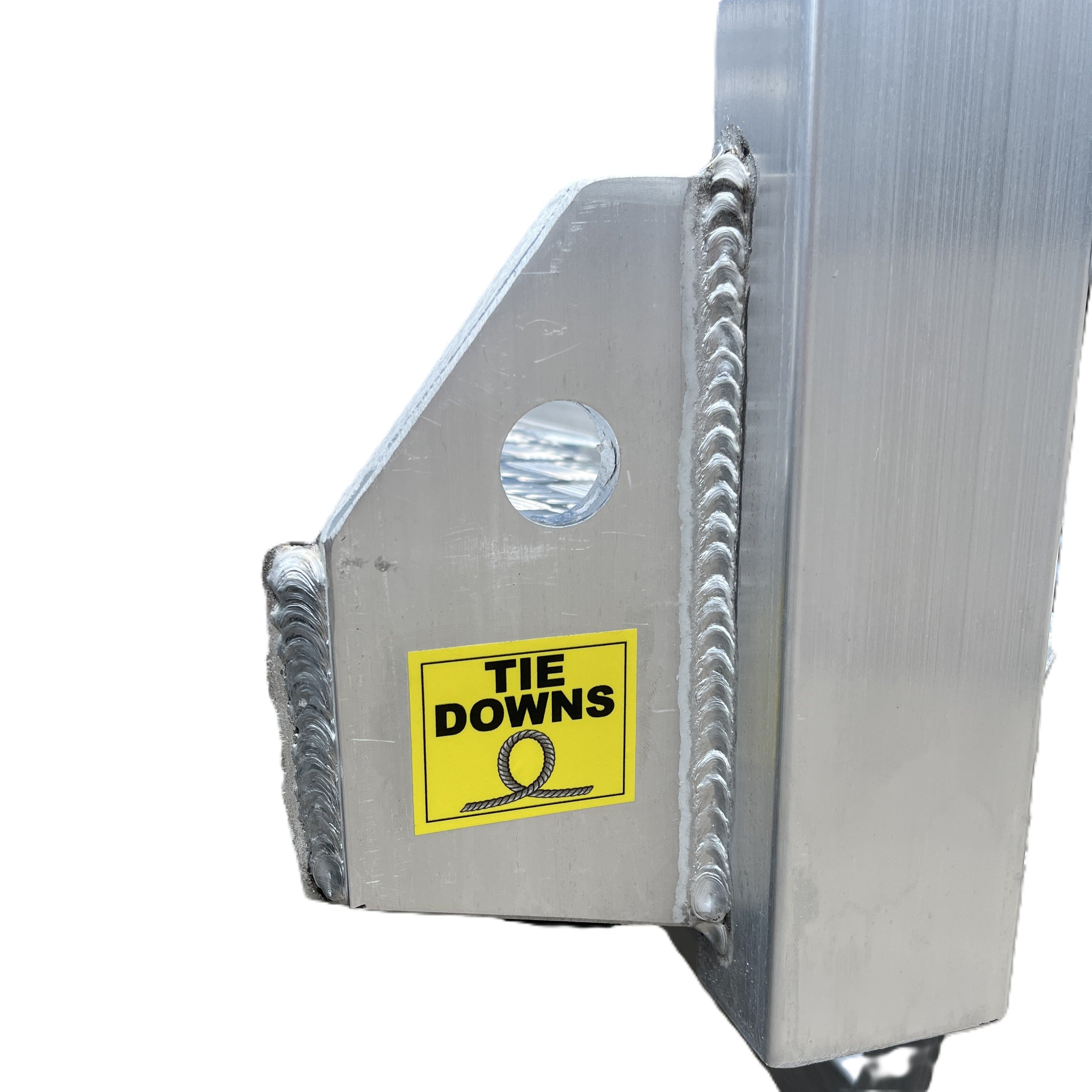 Close-up of a metal structure with a yellow TIE DOWNS label and looped rope graphic on a Paddock Mini Loader & Excavator Alloy Equipment Trailer by Paddock. A blue car is parked in the background on concrete.