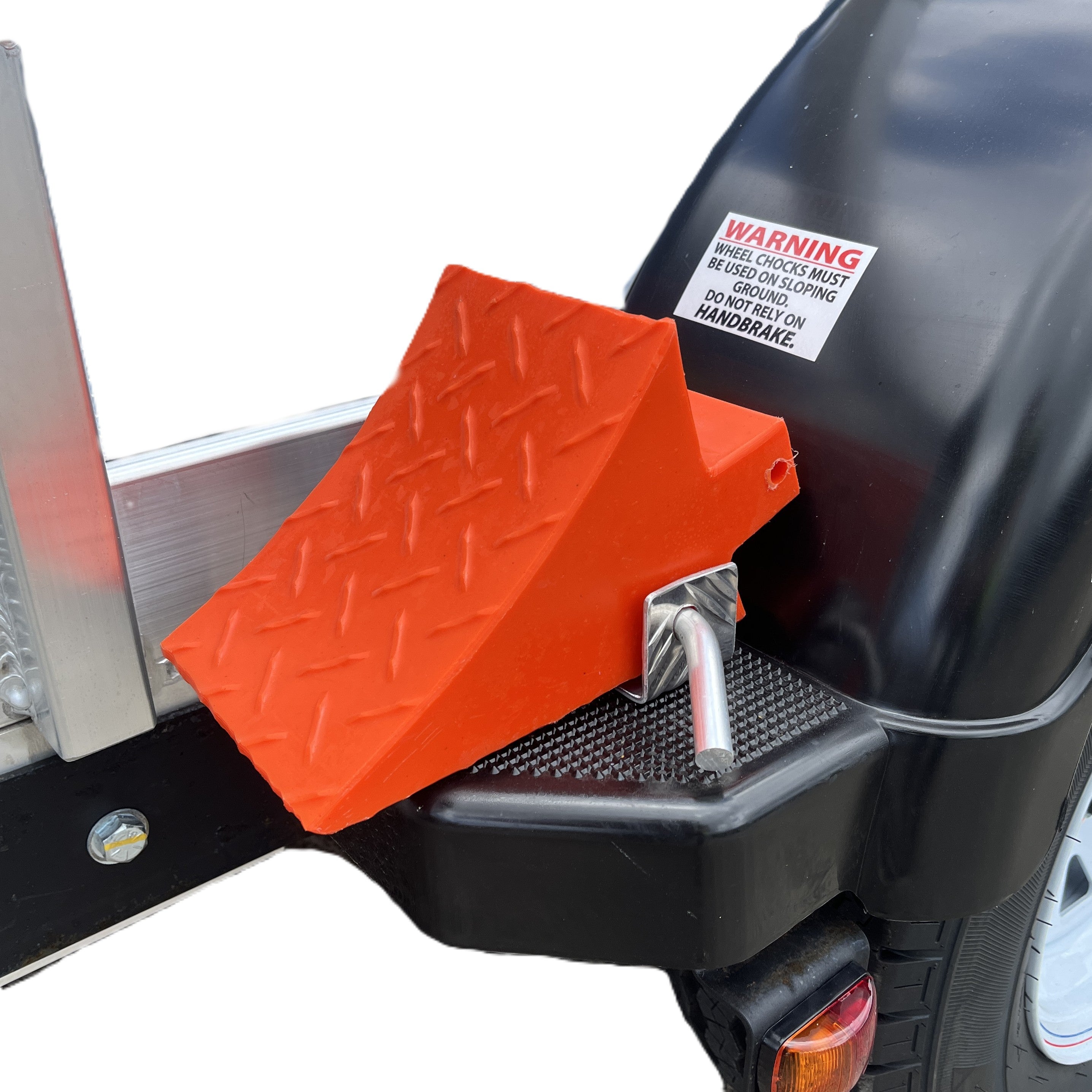 An orange wheel chock with a diamond plate pattern is secured to the black fender of a Paddock Mini Loader & Excavator Alloy Equipment Trailer by a metal pin. A visible warning label and part of a tire can be seen to the right. Brand: Paddock.