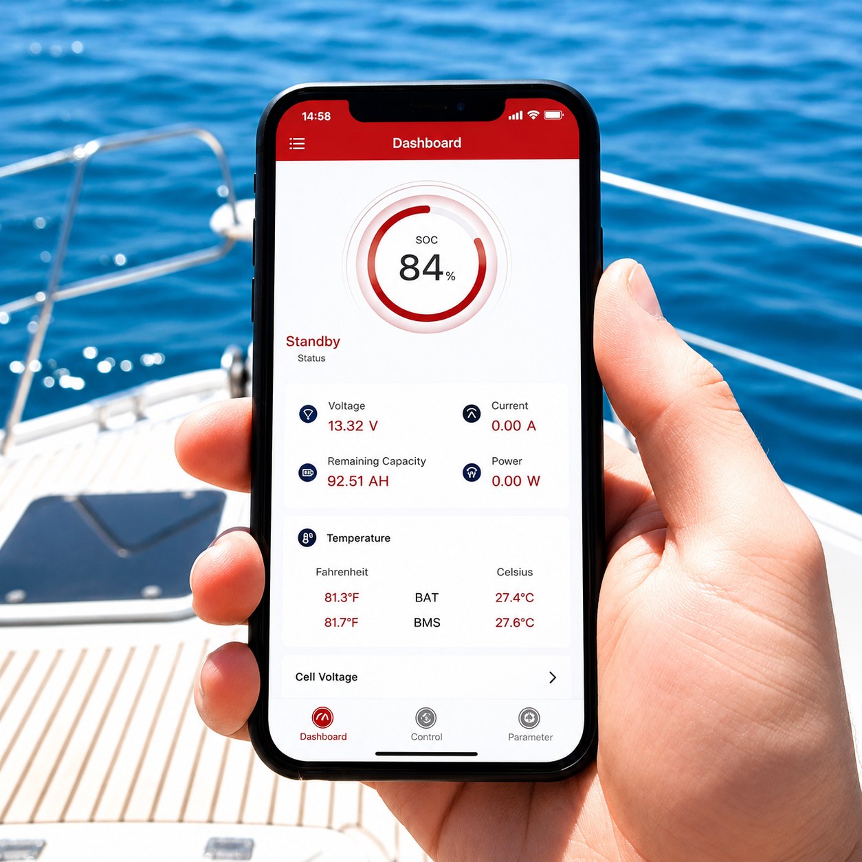 A hand holds a smartphone displaying a battery dashboard app, showing 84% charge on the Trolltek™ 12V LiFePO4 Lithium Slimline Battery AC Bundle—ideal for monitoring your trolling motor’s power—aboard a boat with clear blue water and deck in the background.