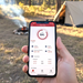 A hand holds a smartphone outdoors near a tent and campfire, with trees and grass in the background. The phone displays the Trolltek Bluetooth app dashboard, showing an 84% charge and technical details—perfect for monitoring your Trolltek™ 12V LiFePO4 Super Slimline Lithium Battery during off-grid adventures.