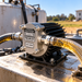 A TurboWerx Exa-Pump TWX-300 high temperature oil scavenge pump with black cooling fins, connected to two clear hoses filled with amber-colored liquid.