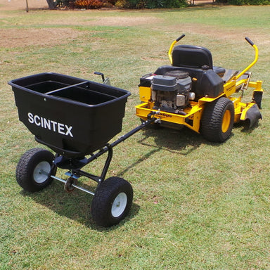 A yellow ride-on lawn mower effortlessly moves over the green grass, towing a black Scintex Seed & Fertiliser Spreader. This tow-behind model with two wheels and a large open container efficiently distributes material under the radiant sun of a clear outdoor day.