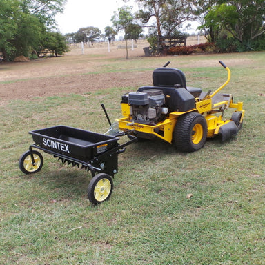 A yellow riding lawn mower is positioned on a grassy field, featuring a black trailer attachment from Scintex known for its Drop Seeder & Spreader. Trees and a fence create the backdrop of the scene.