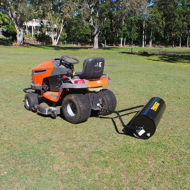 A Scintex Paddock Lawn Roller is being towed by a ride-on lawn mower parked on a grassy field. The heavy-duty steel construction of the roller gleams in the sunlight, casting shadows across the grass. Trees and a fence line can be seen in the background, completing this tranquil scene.