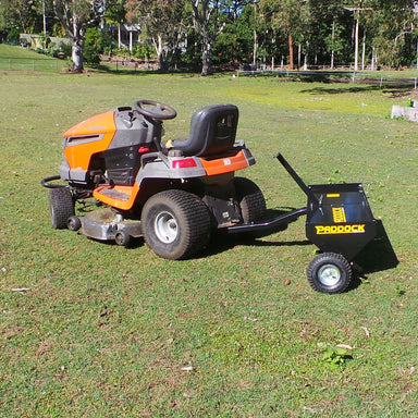 A riding lawn mower with an orange and gray body is parked on a grassy field, accompanied by a Scintex Plug Lawn Aerator. The black Paddock trailer, complete with two wheels, makes hauling the aerator effortless. Trees and a wooden fence offer a tranquil setting, ideal for promoting healthy grass roots.